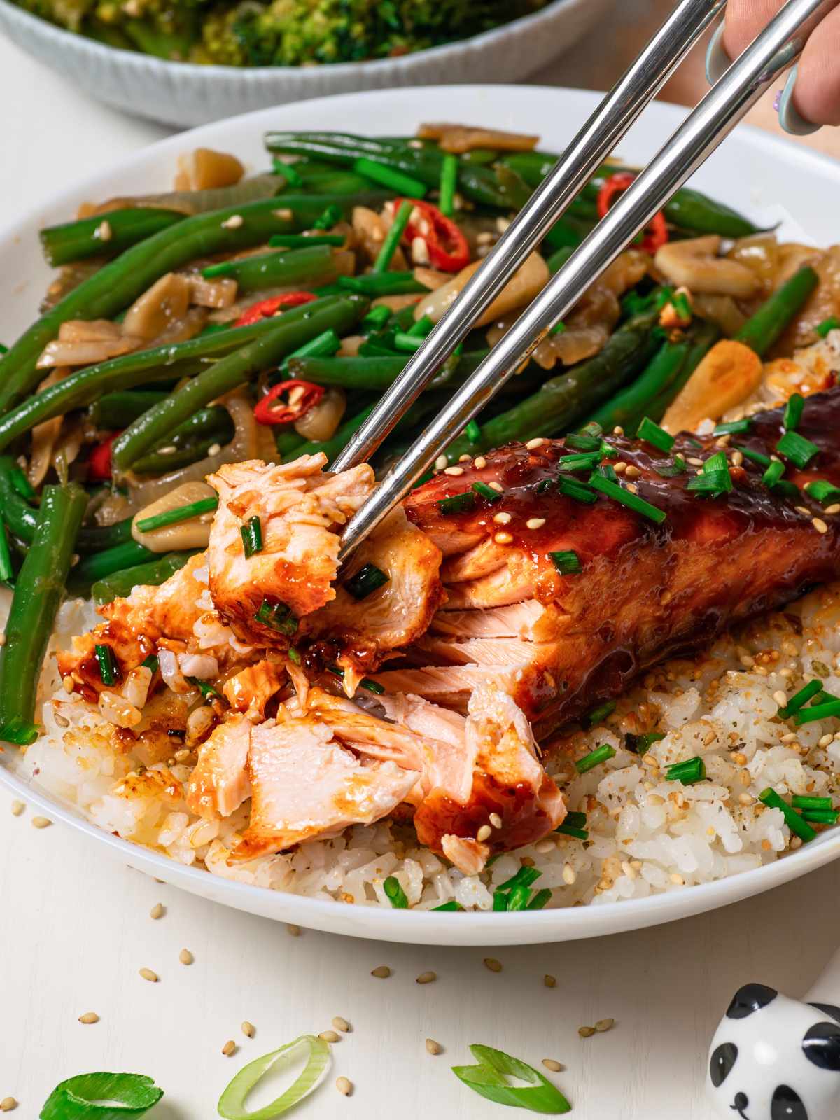closeup of chopsticks holding gochujang salmon on rice with green beans and broccoli on the side
