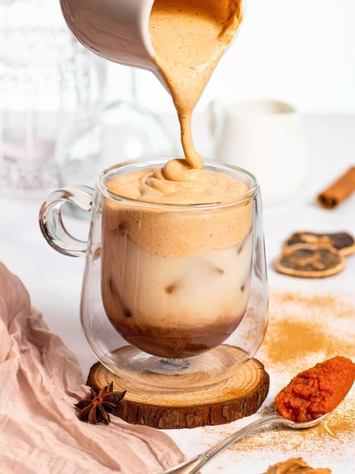 Pouring pumpkin spice cream on top of chai latte in glass