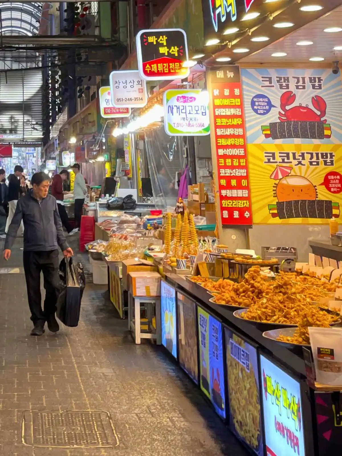 Korean street food stalls in a covered market alley, trays of fried snacks under bright neon menu signs